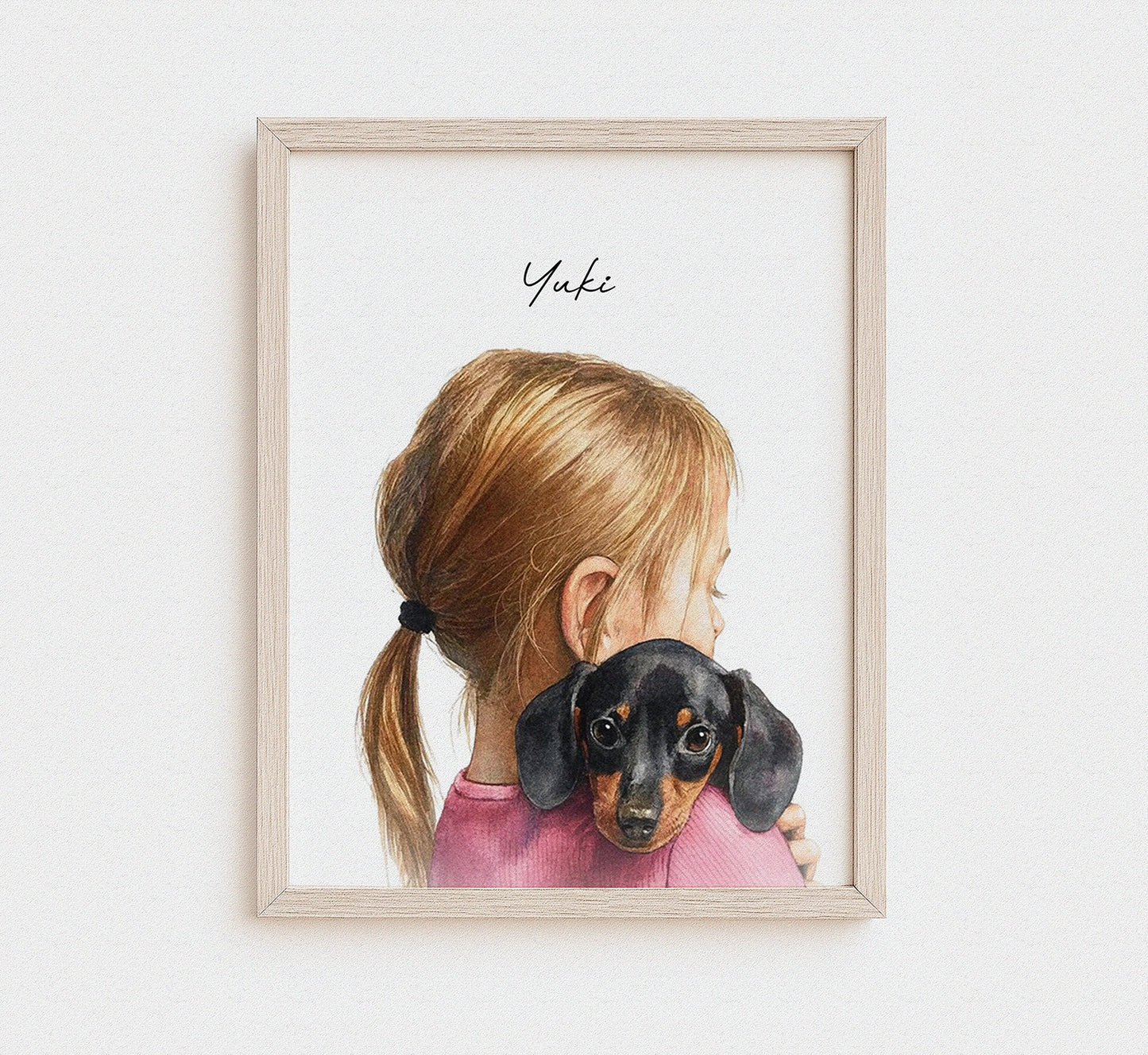 A framed watercolor art print featuring a young girl with blonde hair in a ponytail hugging a black and tan dachshund, with the name 'Yuki' written in script at the top. The print is housed in a light natural wood frame and displayed against a clean white wall.