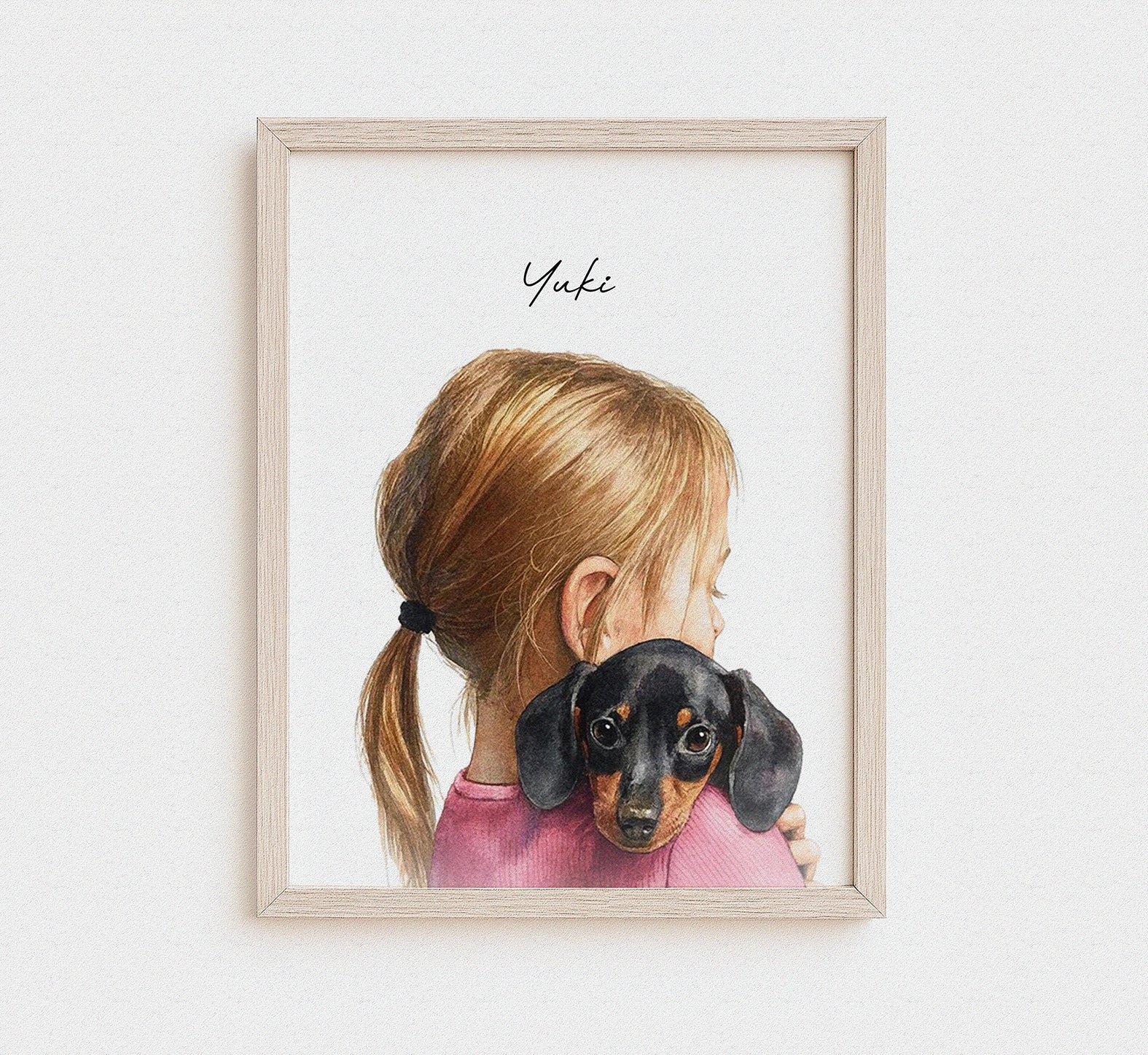 A framed watercolor art print featuring a young girl with blonde hair in a ponytail hugging a black and tan dachshund, with the name 'Yuki' written in script at the top. The print is housed in a light natural wood frame and displayed against a clean white wall.