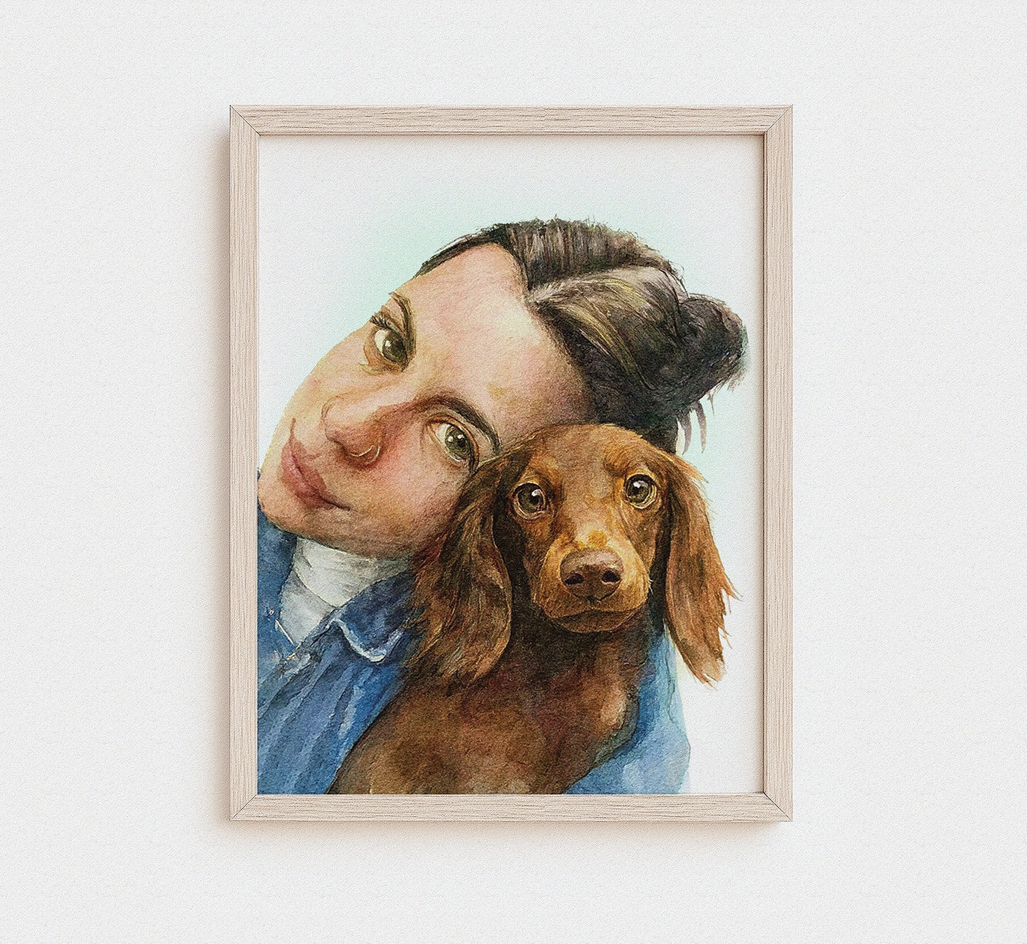A framed watercolor portrait print of a woman hugging a brown dachshund, displayed on a clean white wall.