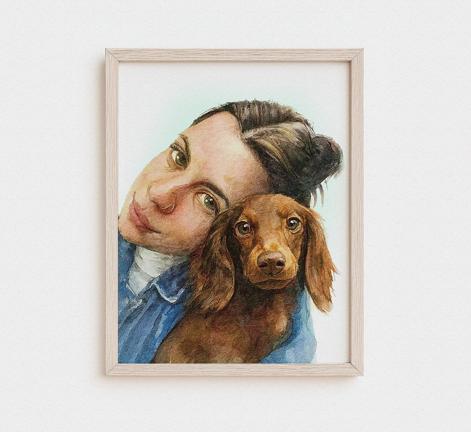 A framed watercolor portrait print of a woman hugging a brown dachshund, displayed on a clean white wall.